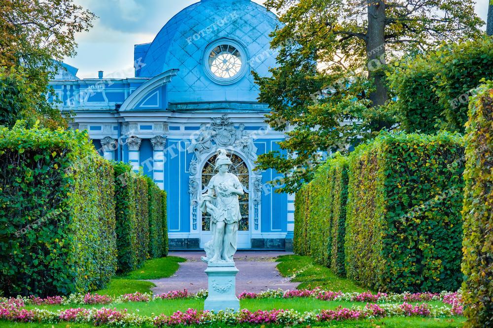 На фотографии изображен Грот в Екатерининском парке Царского Села, построенный в стиле барокко. Здание выкрашено в насыщенные голубые и белые тона, которые гармонично сочетаются с зеленью окружающих аллей и клумб. Перед Гротом стоит классическая статуя, которая добавляет пейзажу историческую элегантность. Этот уголок парка является одним из любимых мест для прогулок и фотографий, привлекая посетителей своей архитектурной изысканностью и живописным оформлением.